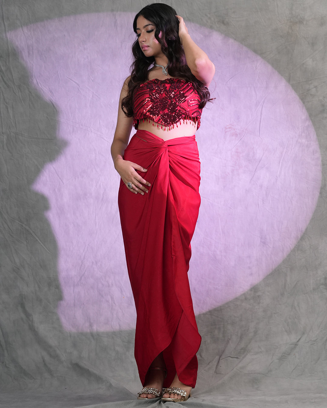 A woman modeling a red two-piece traditional outfit consisting of a strapless embellished crop top and a matching draped skirt, standing against a gray backdrop with a purple spotlight effect.