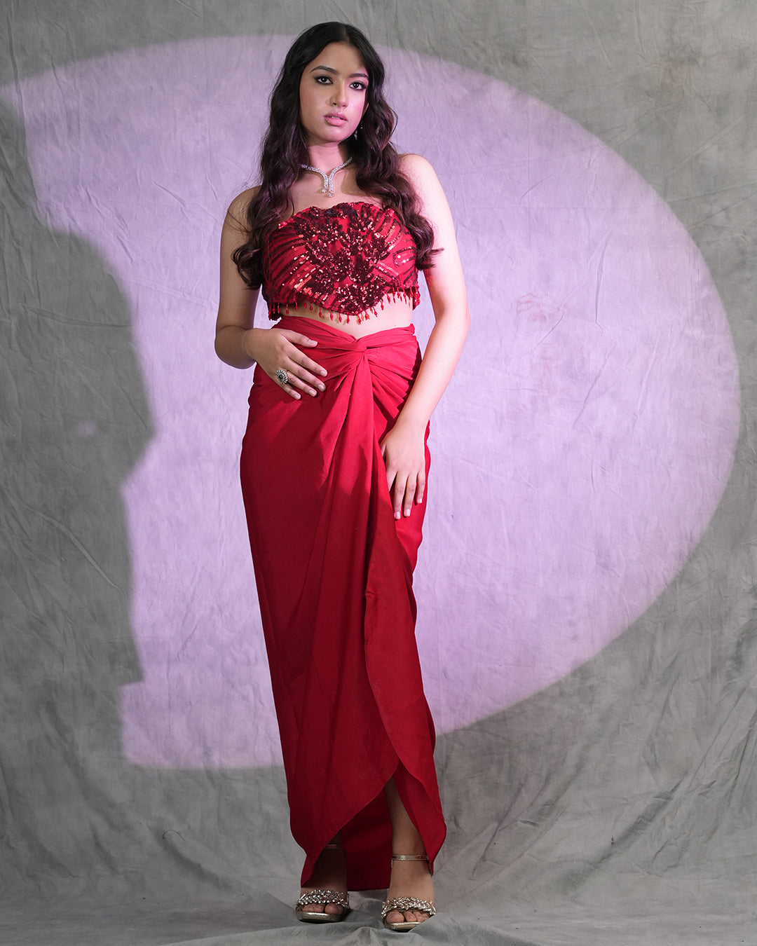 A woman modeling a red two-piece traditional outfit consisting of a heavily embellished red crop top and a matching red draped skirt, standing against a textured gray backdrop with a purple spotlight effect.