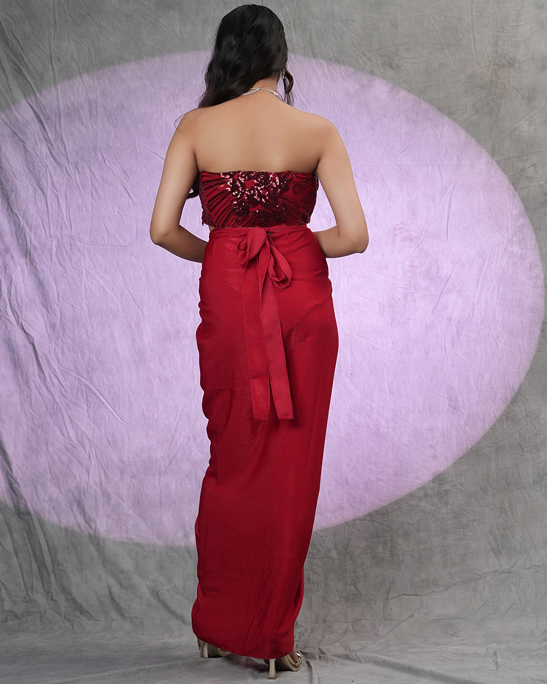 A woman stands with her back to the camera wearing a strapless red gown with a heavily embellished beaded bodice and a large bow at the back waist, set against a gray backdrop with a large lavender spotlight effect.