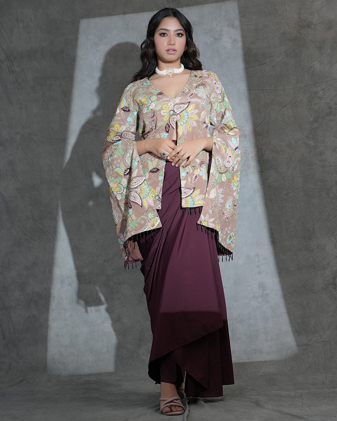 A woman models a traditional outfit featuring a beige and multicolor floral printed shawl draped over a deep maroon dress, set against a textured gray studio backdrop with dramatic lighting.
