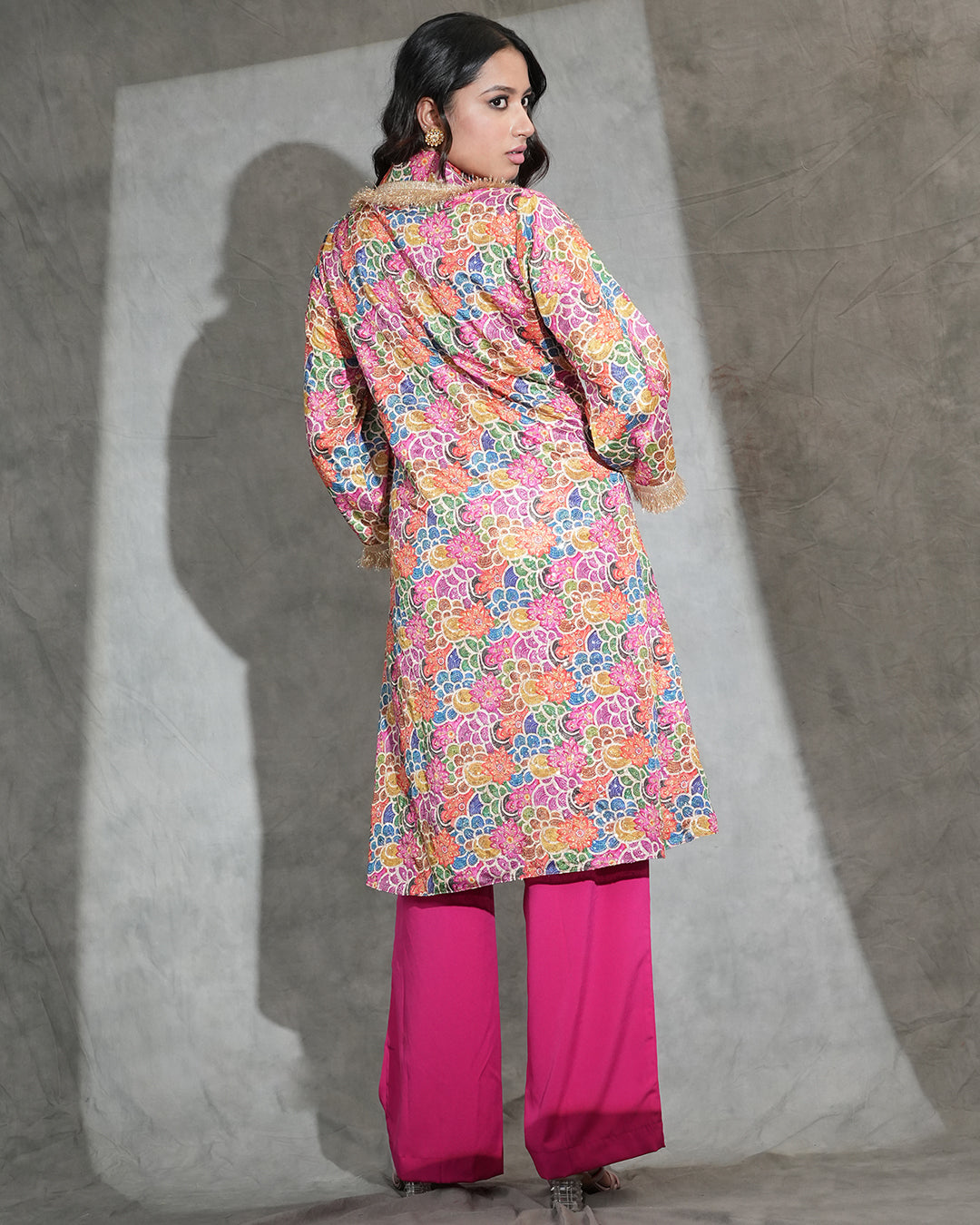 A woman stands with her back to the camera wearing a long-sleeved, multicolor floral printed tunic with a fuzzy collar, paired with bright pink wide-leg pants, against a textured gray wall with a spotlight.