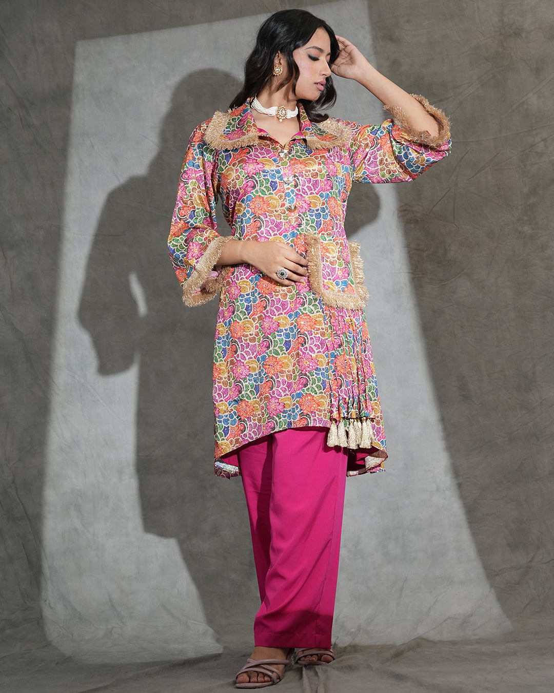 A woman models a vibrant, multicolored floral printed kurta with beige faux-fur trim and tassel details, paired with bright pink pants, against a textured gray studio backdrop.