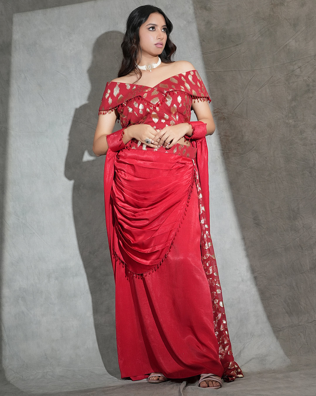 A woman modeling a red off-the-shoulder saree with gold geometric patterns and a matching red dupatta, standing against a textured gray studio backdrop.