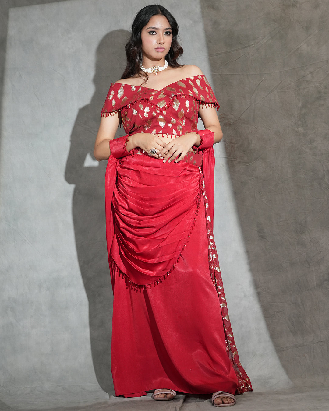 A woman modeling a red off-the-shoulder gown with gold floral embroidery on the blouse and border, standing against a textured gray backdrop.