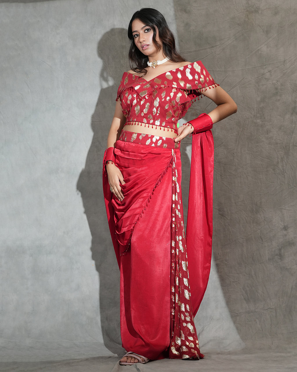 A woman modeling a bright gown with white floral patterns, paired with a matching red and white embroidered blouse, standing against a textured gray studio wall.