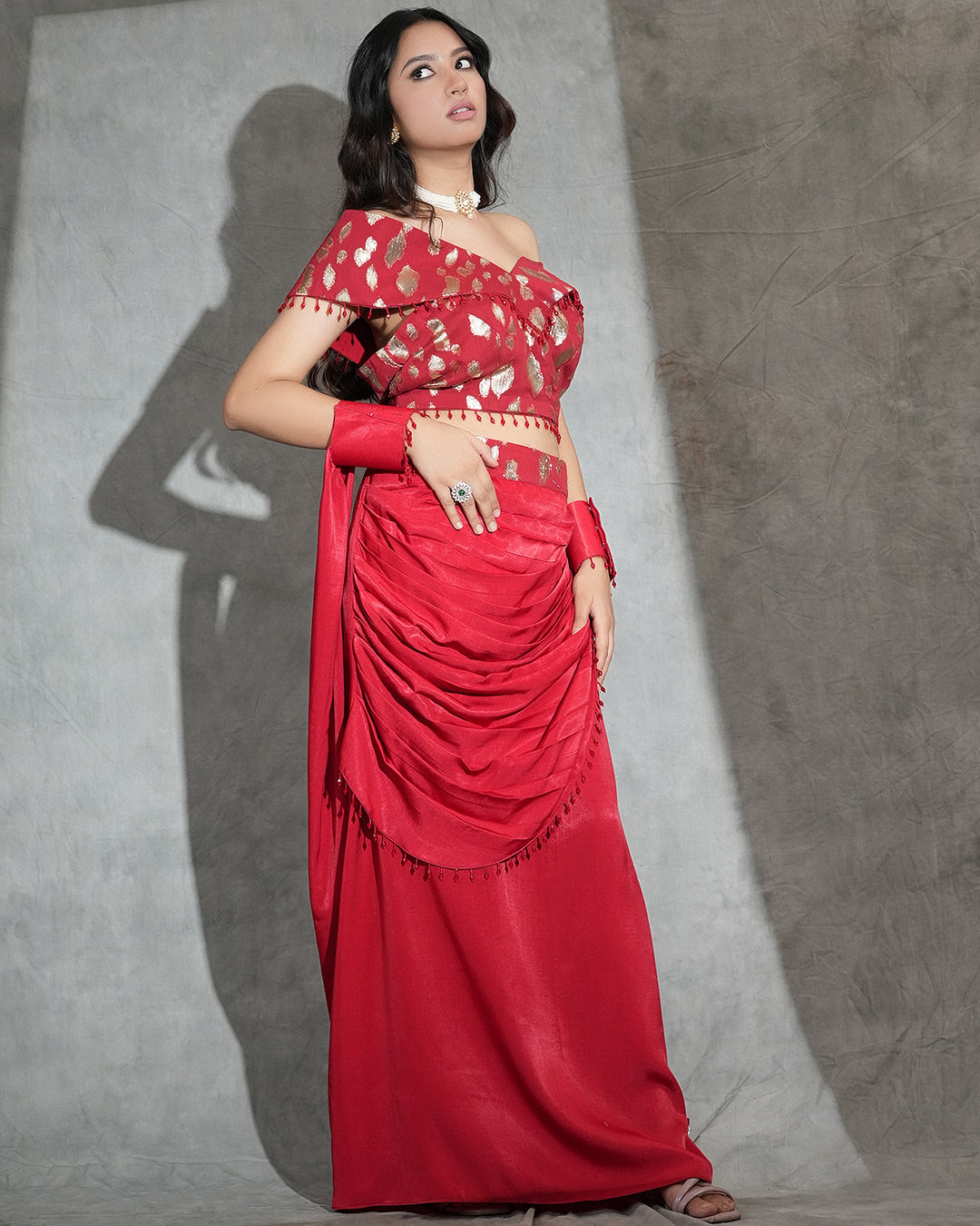 A woman models a red off-the-shoulder saree with gold and white floral patterns on the blouse and a matching draped skirt, posed against a textured gray backdrop with a strong shadow.