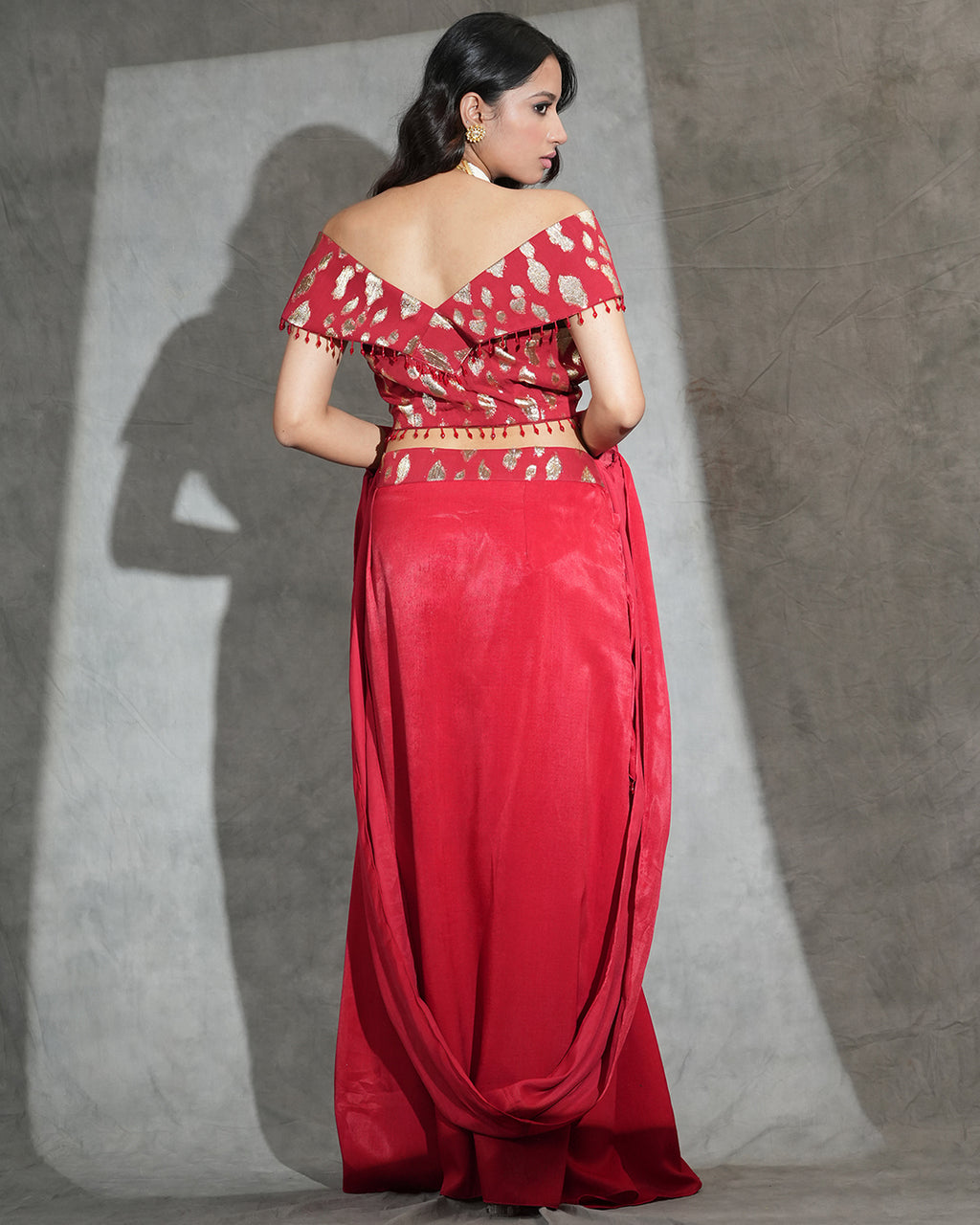 A woman modeling a red off-the-shoulder maxi dress with gold floral embroidery and a matching embroidered waistband, shown from the back against a textured gray backdrop.