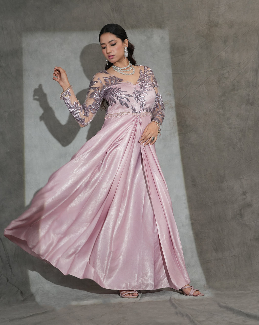 A woman modeling a floor-length pink gown with sheer embroidered sleeves and a flowing satin skirt, posed against a textured gray backdrop.