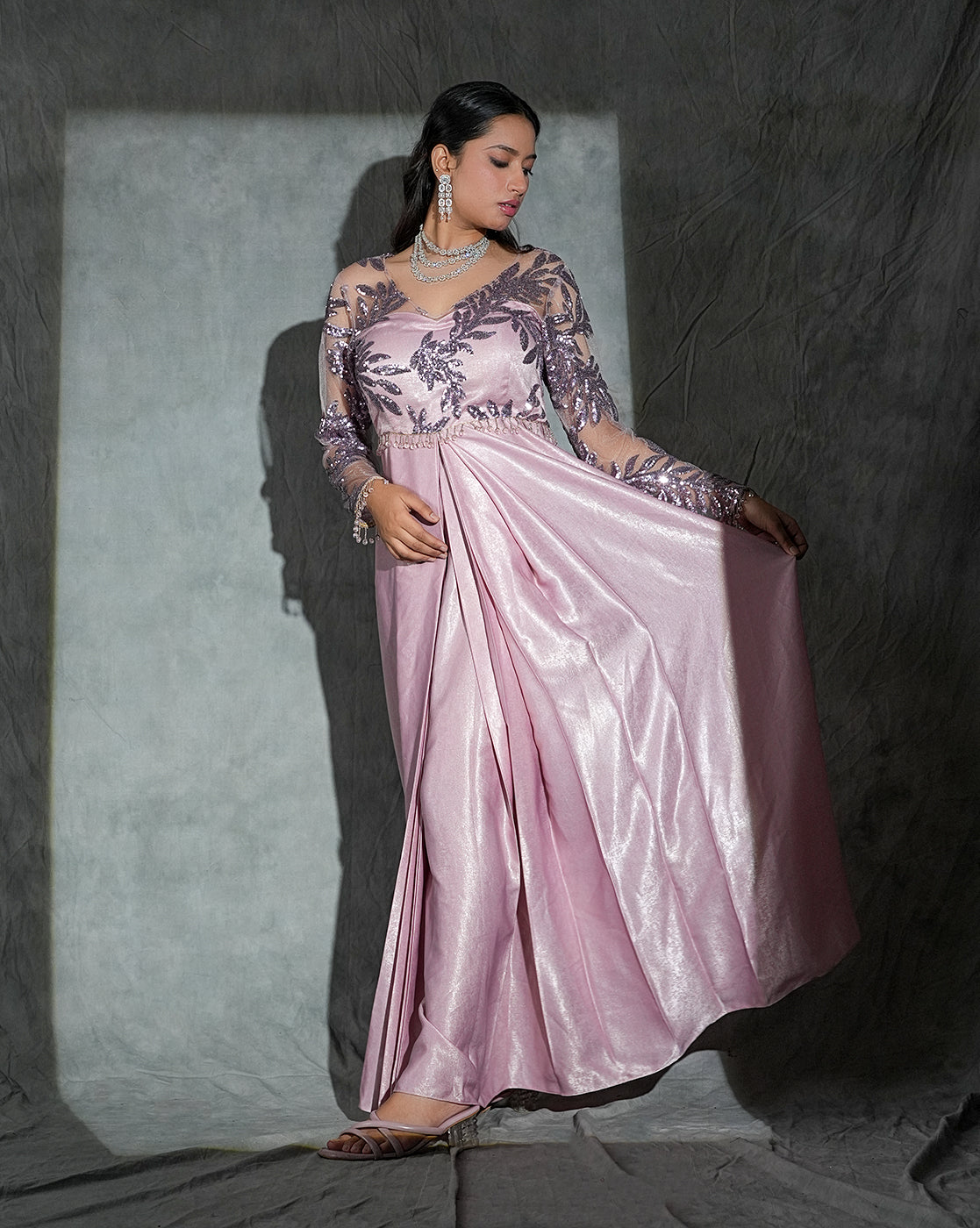 A woman models a traditional Indian salwar kameez gown in a dusty pink color. The dress features a sheer floral embroidered bodice with long sleeves and a flowing satin skirt, posed against a textured gray backdrop with dramatic lighting.