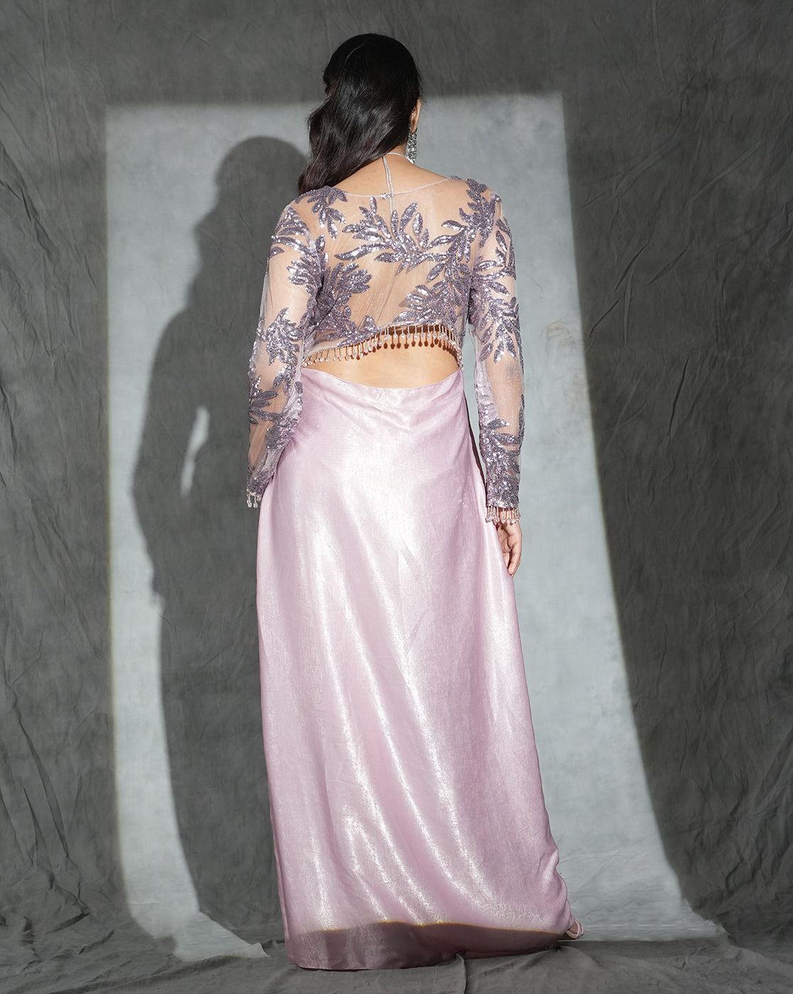 A woman shown from the back wearing a floor-length formal gown with a shimmering pale pink skirt and a sheer embroidered top in lavender-purple tones, posed against a textured gray backdrop with a spotlight behind her.